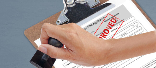 Close up of hand holding a rubber stamp above Land Use Application with "approved" stamped in red on the application