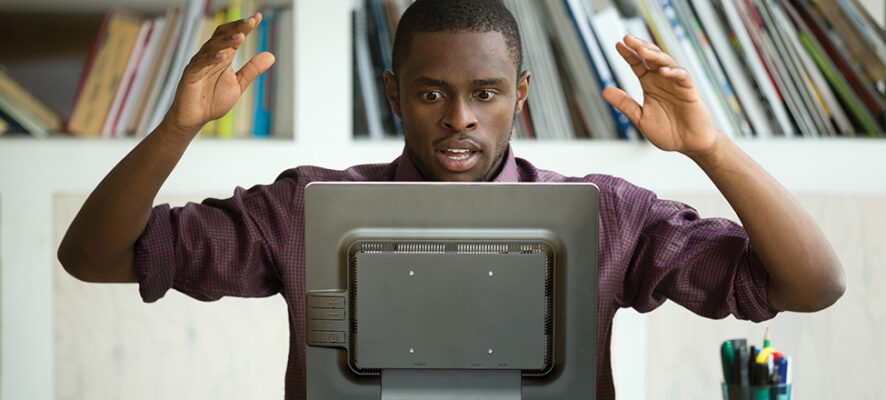 African American man raises hands in disbelief as looks at computer monitor in negative surprise