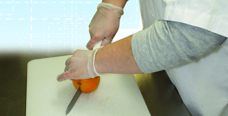 Close up on food service worker wearing gloves cutting orange in half on cutting board against a light blue background
