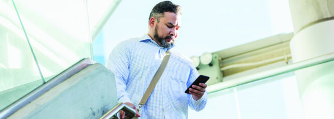 A mid adult Hispanic man in his 30s wearing a button down shirt, walking down stairs outside a building, looking down at his mobile phone.
