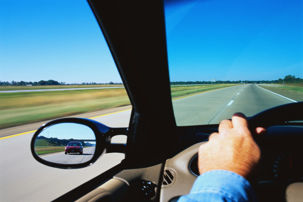 Driver's perspective while driving on a highway: close up of side mirror, hand on steering wheel and view from windshield and side window