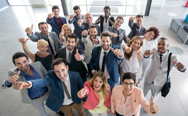 Group of business people in lobby smile at camera each with thumbs up gesture