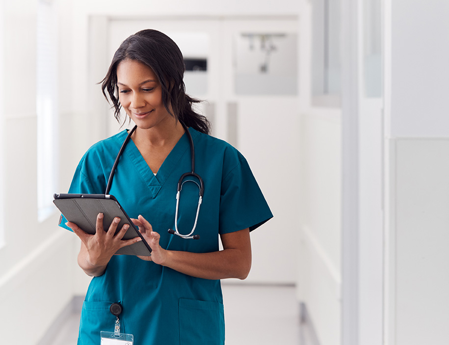 Female Doctor Wearing Scrubs In Hospital Corridor Using Digital Tablet