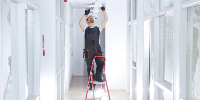 Maintenance worker stands on step stool to work on overhead light fixture.