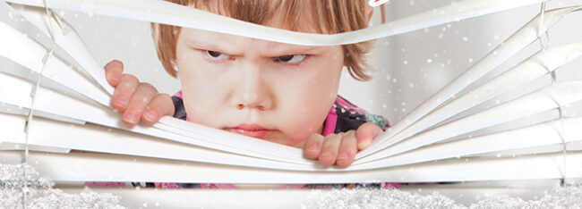 Young girl with scowl on face looks through blinds at snow falling