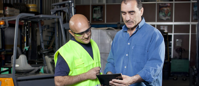 Two men wearing safety gear working with a forklift. One man showing the other information on a digital tablet.