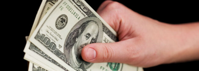 Hand holds stack of $100 bills fanned out against a black background.