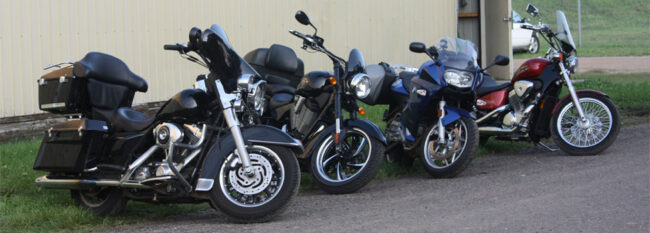 Row of four motorcycles parked along dirt road. Two black motorcycles and a blue and red motorcycle.