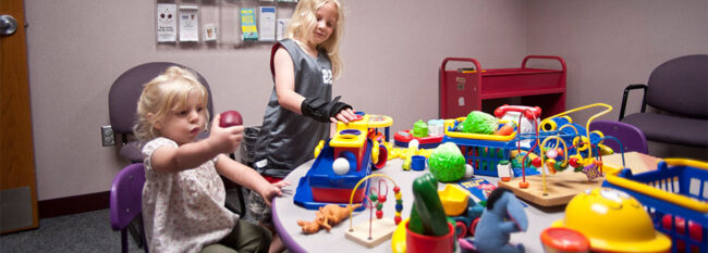 Two children playing with toys in doctors waiting room. One child is wearing a grey 22 jersey. The other is wearing a pink floral shirt.