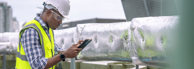 Man inspecting industrial air conditioning unit. Taking notes on tablet. Wearing white helmet, safety glasses, yellow vest and blue plaid shirt.