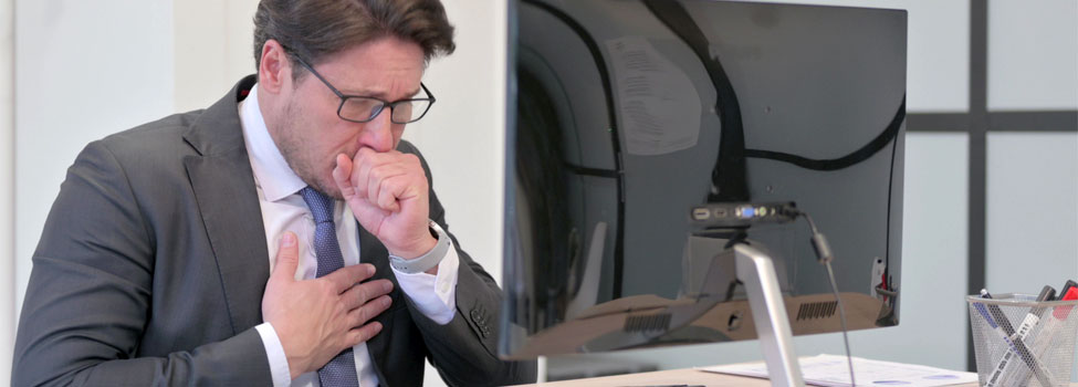 Man in suit sitting at desk coughing and grabbing chest in front of computer.