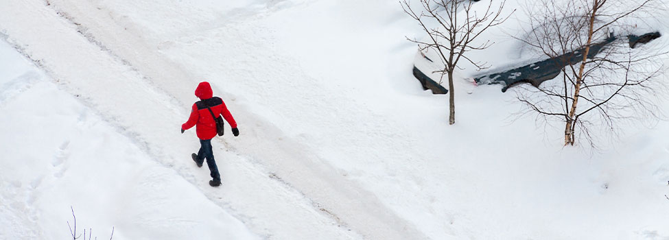 Man walking in winter on snow plowed path near parking lot. Man wearing red jacket with shoulder bag.