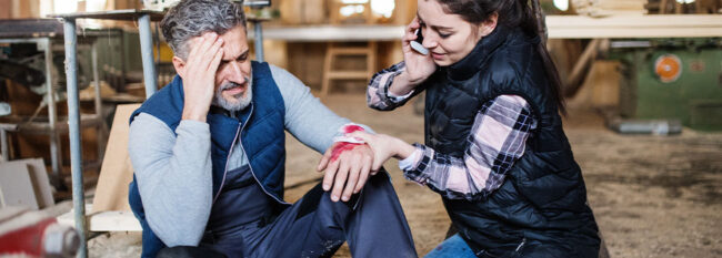 Man has hurt his hand in workshop and holding his other hand to head in pain. Woman is holding a bloody bandage to his hand and calling for help. Both are sitting on the ground.