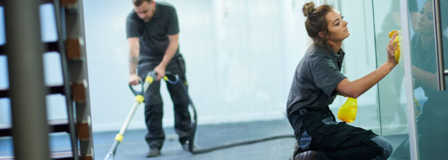 Crew cleaning office. Woman wiping window and man vacuuming. Both focused on their jobs.