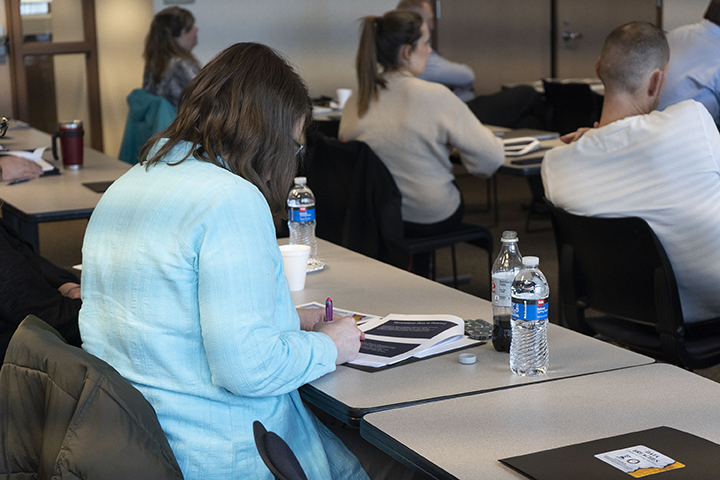 Woman takes notes during a training seminar. Other attendees are attentive to speaker.