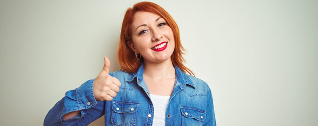 Young beautiful redhead woman wearing denim shirt standing over white isolated background doing happy thumbs up gesture with hand.