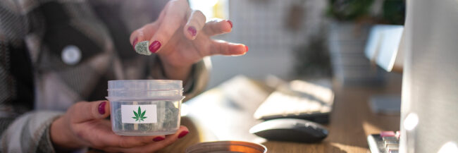 Woman takes small cannabis edible from container while seated at desk