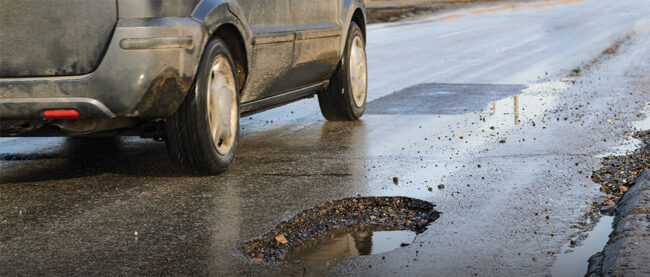 Close up of road with pothole while car drives near it