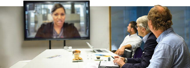 Group of business people gathered in person while one person joins the meeting using video teleconferencing technology
